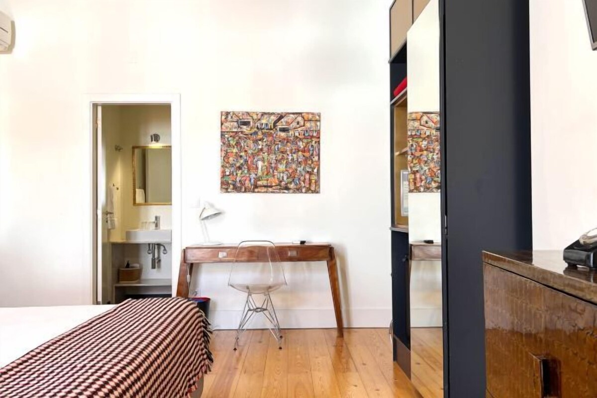 Une chambre avec un lit, un bureau et une chaise. Il y a une porte qui donne sur une salle de bain et un miroir est accroché au mur.