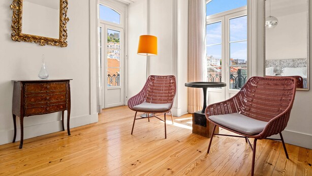 Amplia habitación con piso de madera, un ventanal con vistas a edificios, una silla de mimbre roja, una pequeña mesa con una lámpara y un espejo decorativo.