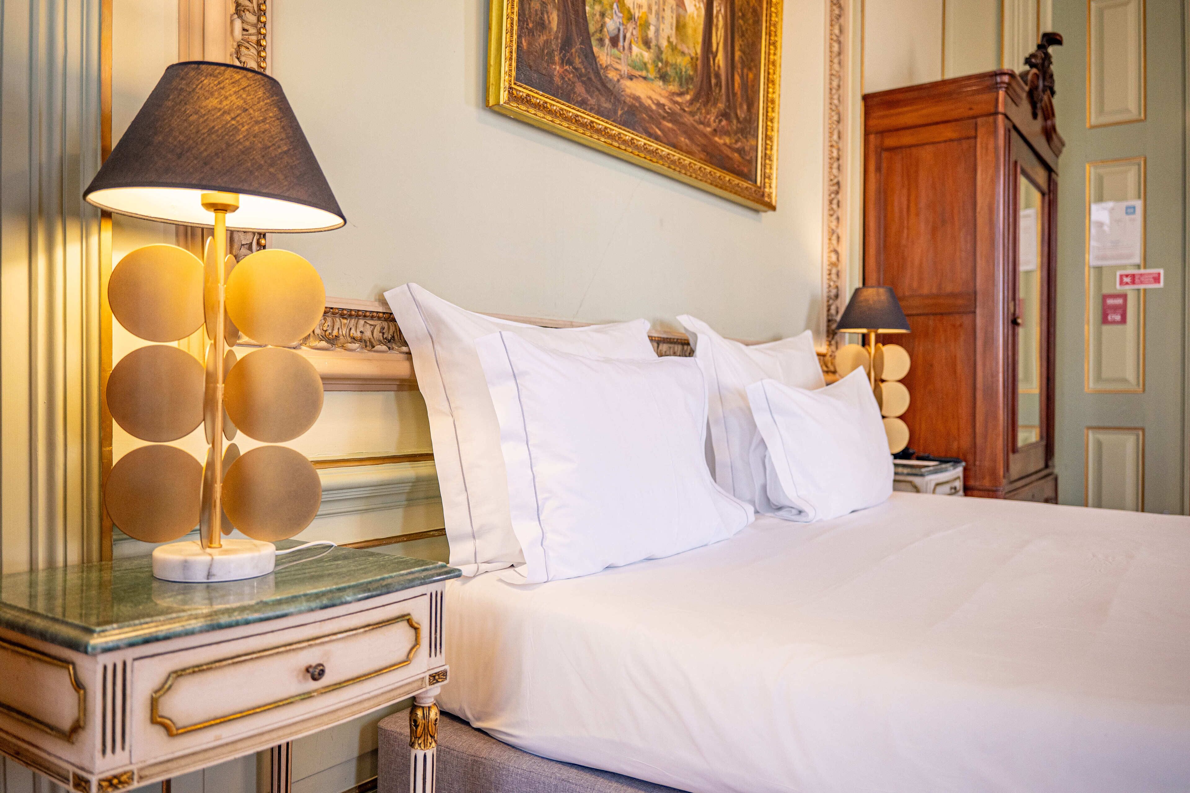A neatly made bed with white linens, a round table with a lamp, and a painting on the wall.