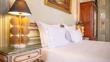 A neatly made bed with white linens, a round table with a lamp, and a painting on the wall.