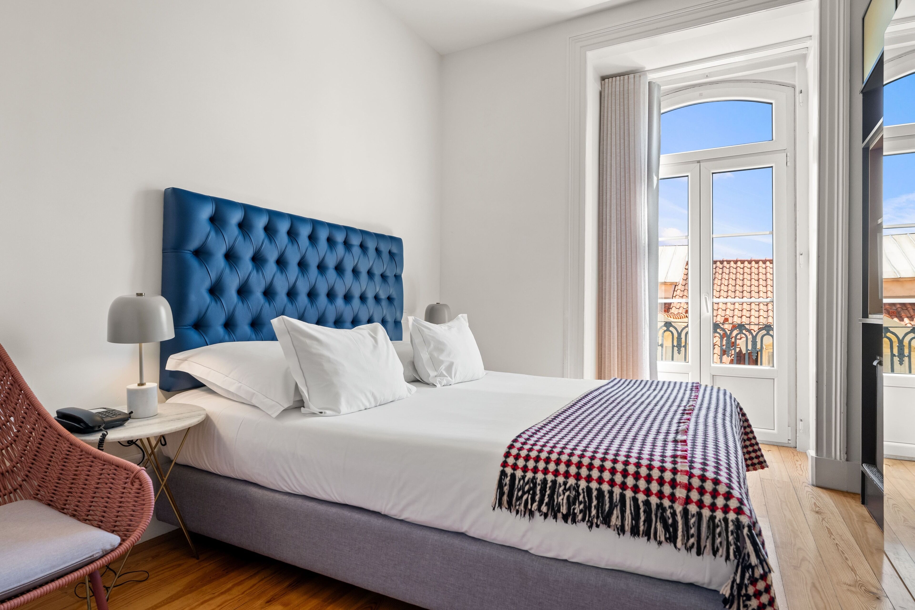A hotel room with a large bed, a blue headboard, white bedding, a patterned throw blanket, a small side table with a lamp, and a wicker chair.