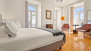 Un dormitorio amplio con una cama grande, una mesita, una silla y vistas a la ciudad a través de amplios ventanales.