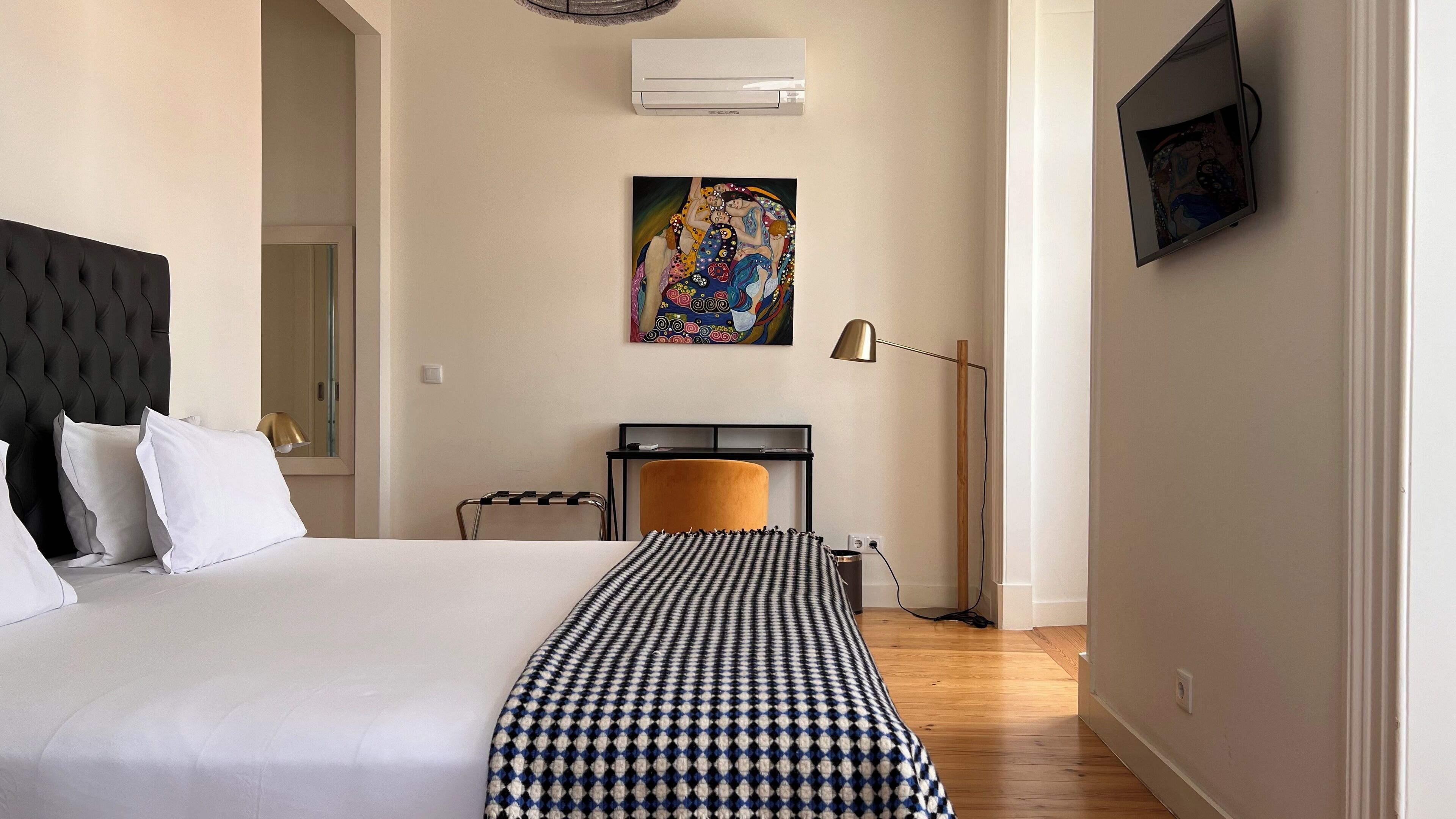 A hotel room with a large bed, a headboard, a painting on the wall, a desk with a chair, and a lamp.