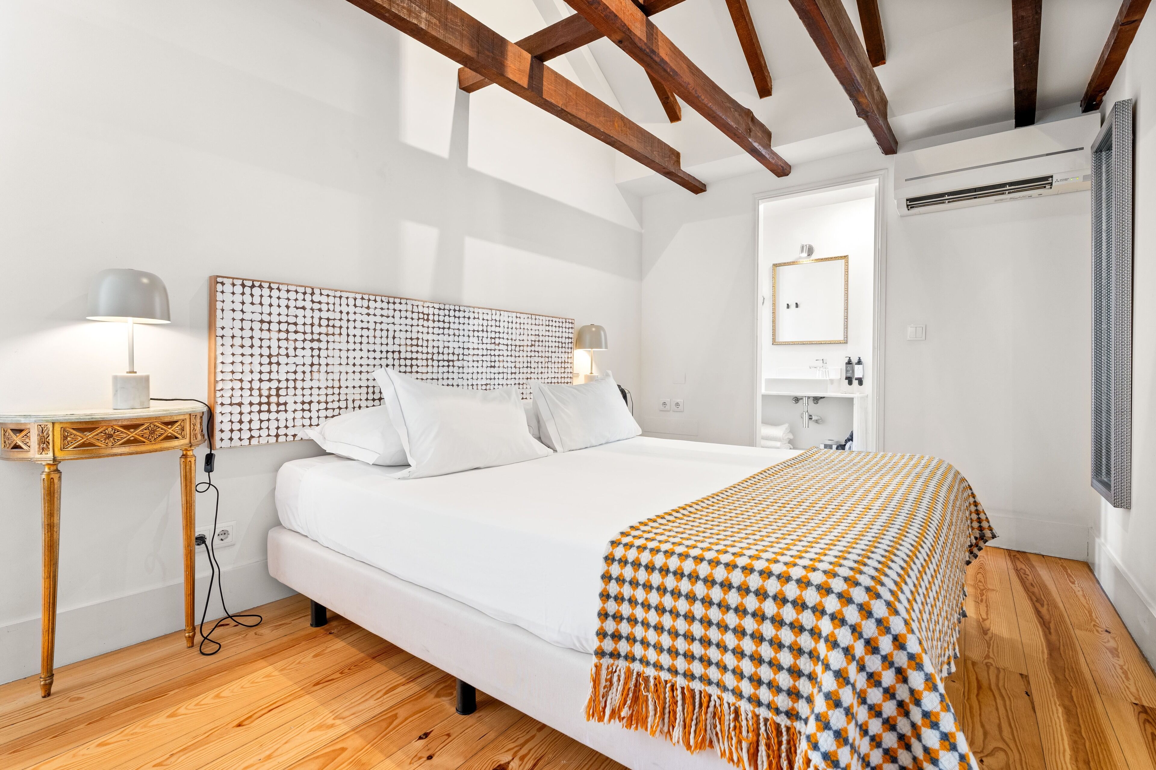 Quarto com uma cama grande, vigas de madeira no teto, mesa de cabeceira com luminárias e um espelho na parede.