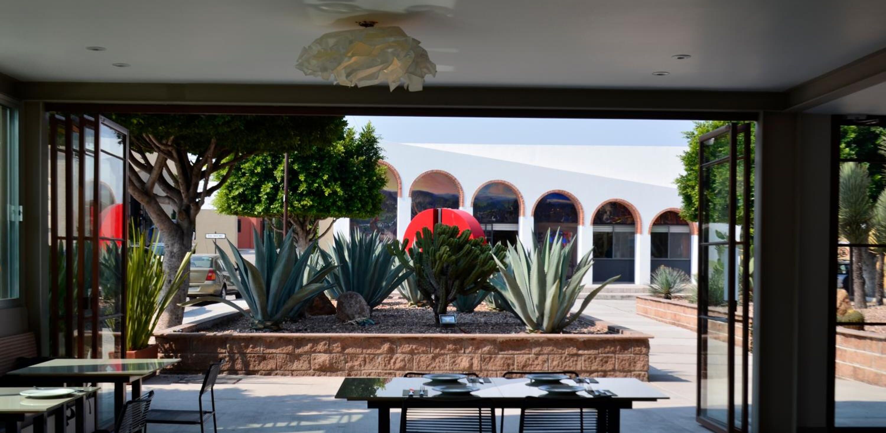 An indoor dining area with tables and chairs, a decorative ceiling light, and a view of a building with arched windows and a red umbrella.
