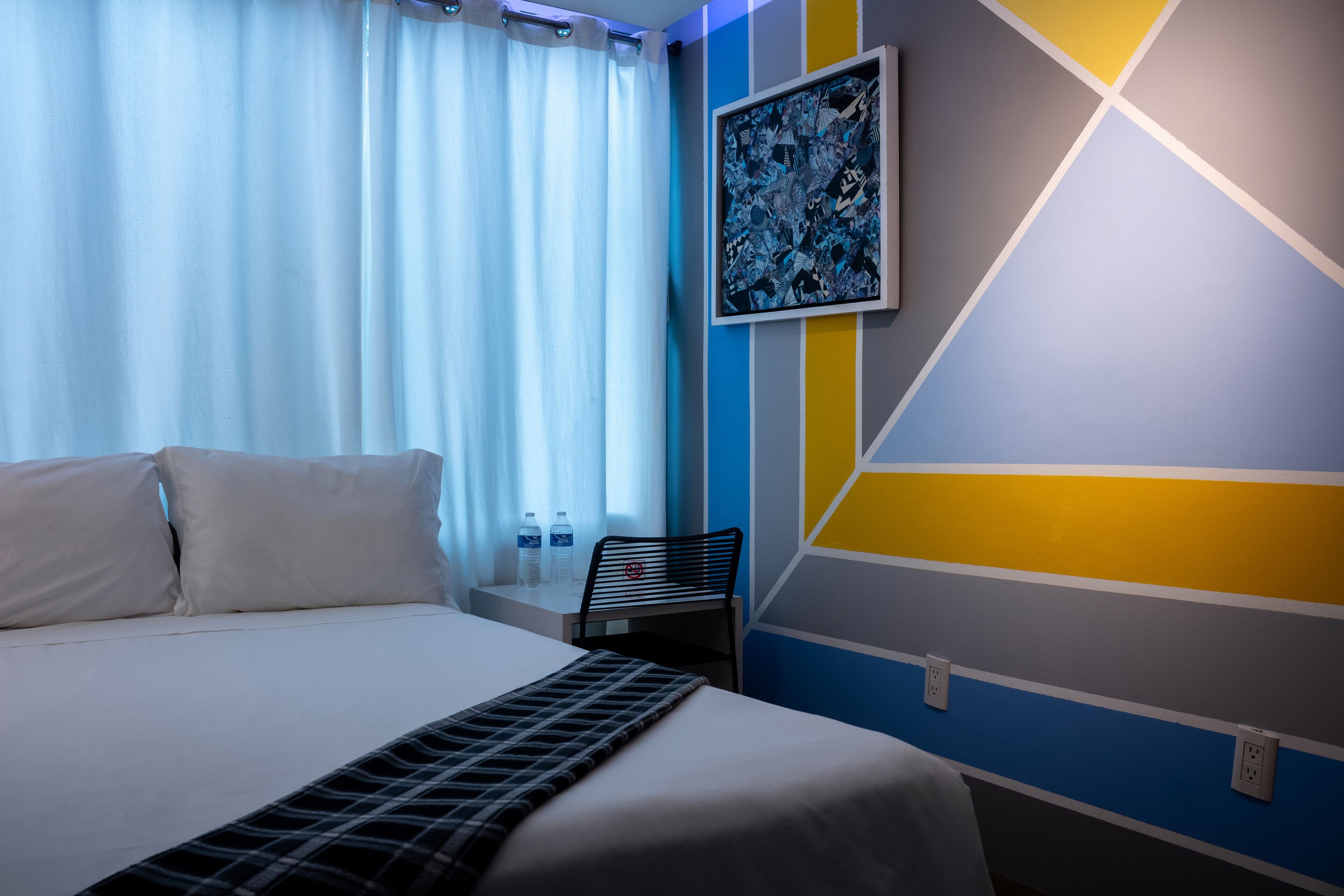 Une chambre d’hôtel avec un lit, une chaise, une table avec des bouteilles d’eau et un mur orné de motifs géométriques.