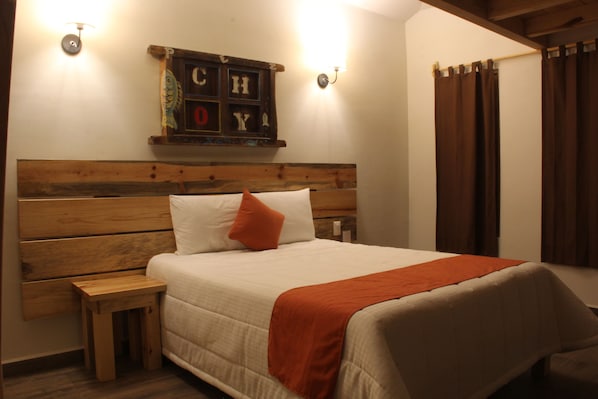 Un dormitorio con cabecera de madera, una mesita de noche pequeña, cama con ropa de cama blanca y un almohadón naranja, y un estante en la pared con adornos.