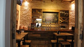 Un rústico área de bar con mesas y taburetes de madera, pared de piedra y un menú en pizarra que anuncia diferentes cervezas.