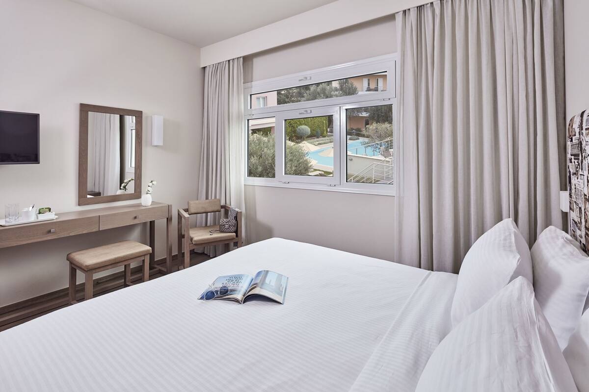 A hotel room with a bed, a desk, a chair, a window with a view of a pool, and a flat-screen TV.