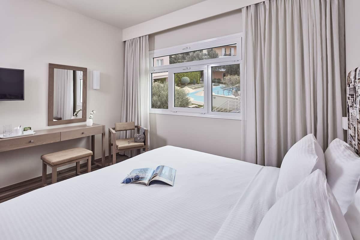 A hotel room with a bed, a desk, a chair, a window with a view of a pool, and a flat-screen TV.