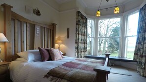 A bedroom with a large bed, two bedside lamps, a window with curtains, and a view of trees outside. - Cormiston Farm (Biggar)