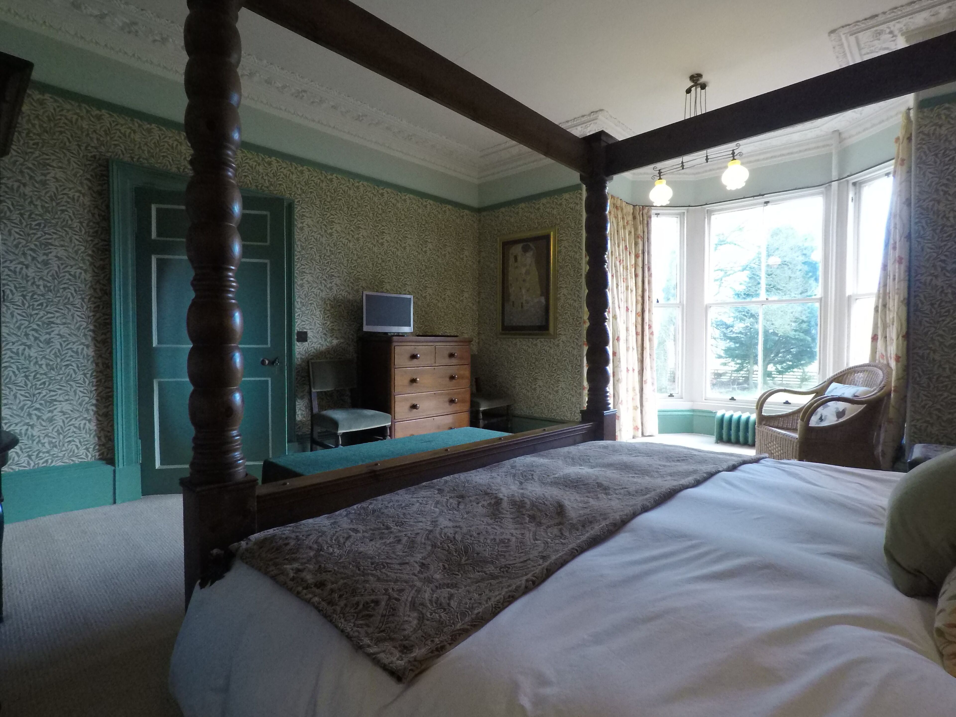 A bedroom with a four-poster bed, a dresser, a television, and a window with a view of trees.