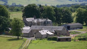 A stone farmhouse with multiple chimneys and a barn, surrounded by lush green fields and trees. - Cormiston Farm (Biggar)