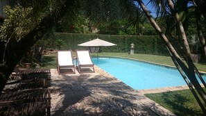 A swimming pool with two sun loungers and a covered umbrella, surrounded by greenery and a stone pathway.
