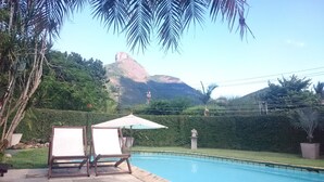 A swimming pool with two lounge chairs and an umbrella, surrounded by lush greenery and a mountain in the background.