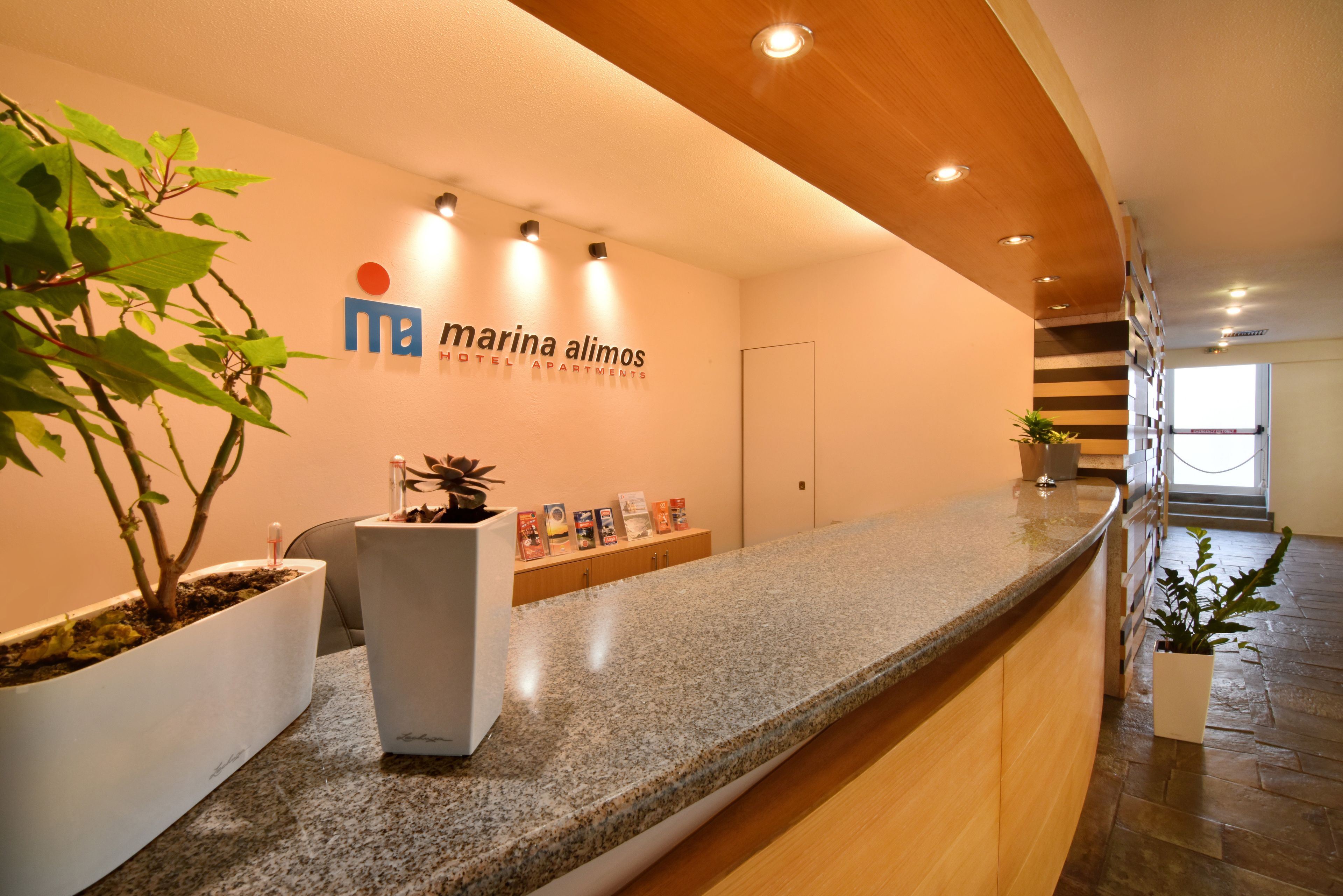 Een receptie met een granieten balie, plantenbakken en een bord met 'Marina Alimos Hotel & Apartments'.
