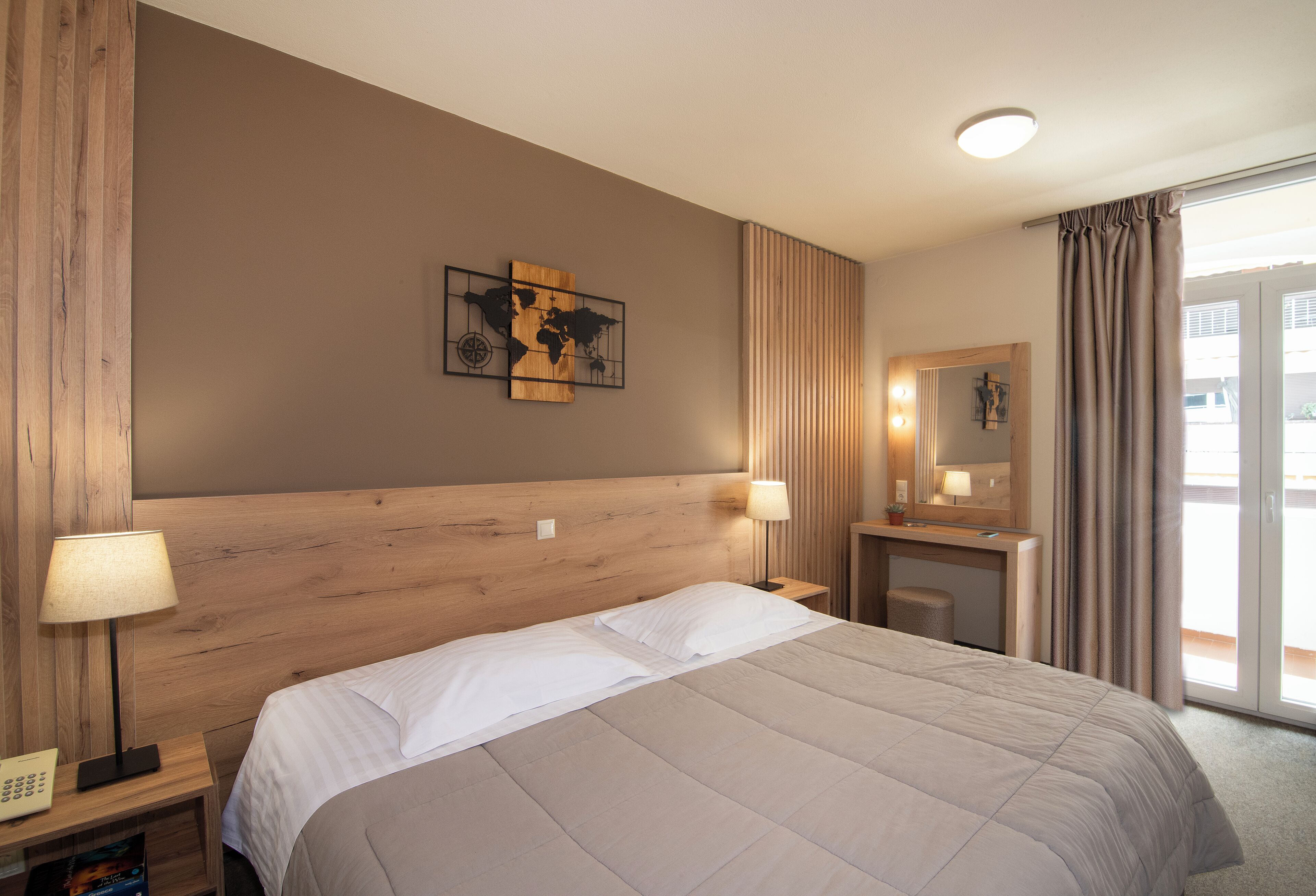 Ein Hotelzimmer mit einem großen Bett, einem Holzkopfstück, zwei Nachttischlampen, einem kleinen Schreibtisch mit Spiegel und einem Wandbild einer Weltkarte.