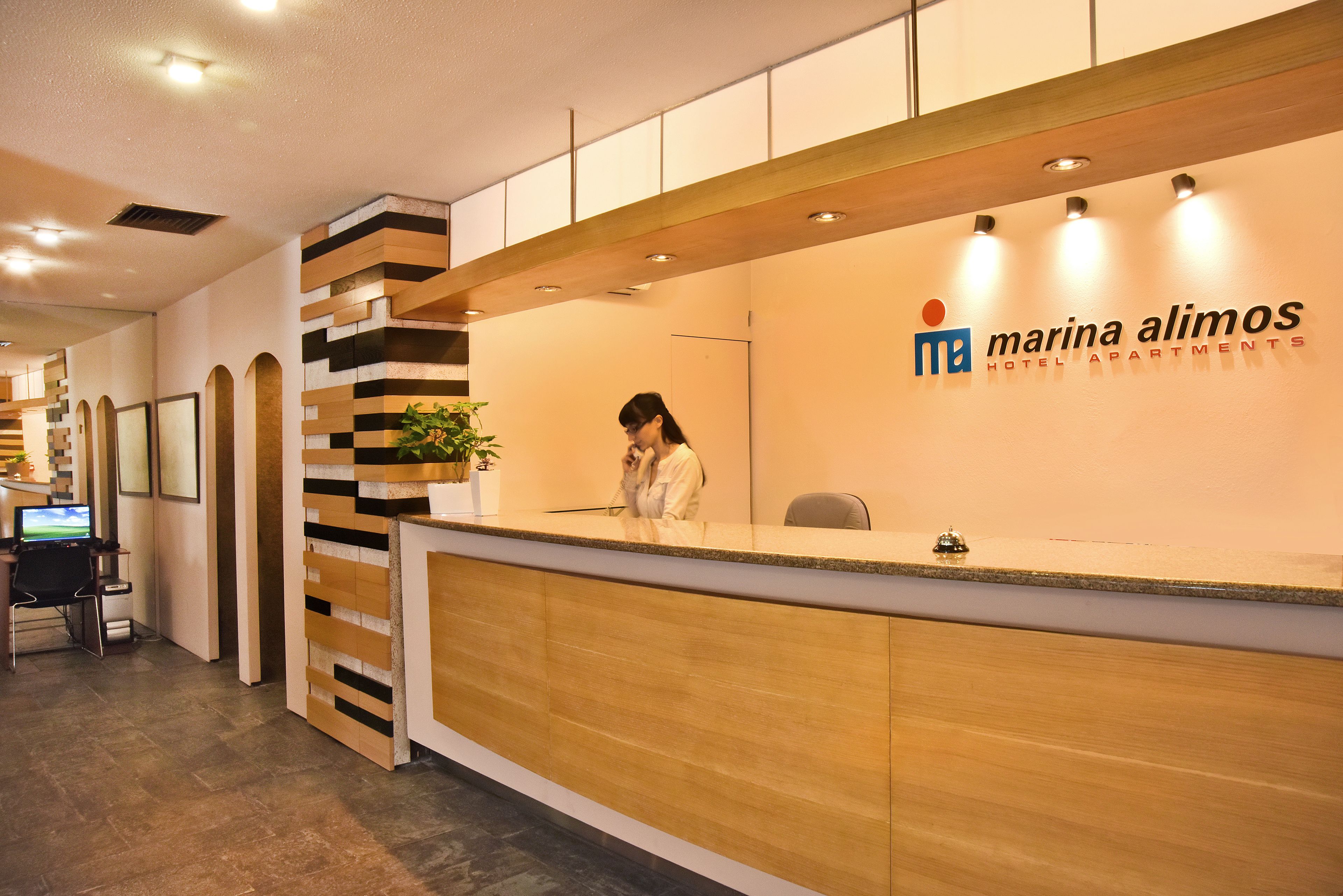 A reception desk with a receptionist, a logo for 'Marina Alimos Hotel Apartments', and a view of the sea through the window.