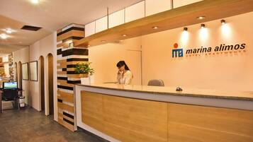 A reception desk with a receptionist, a logo for 'Marina Alimos Hotel Apartments', and a view of the sea through the window.