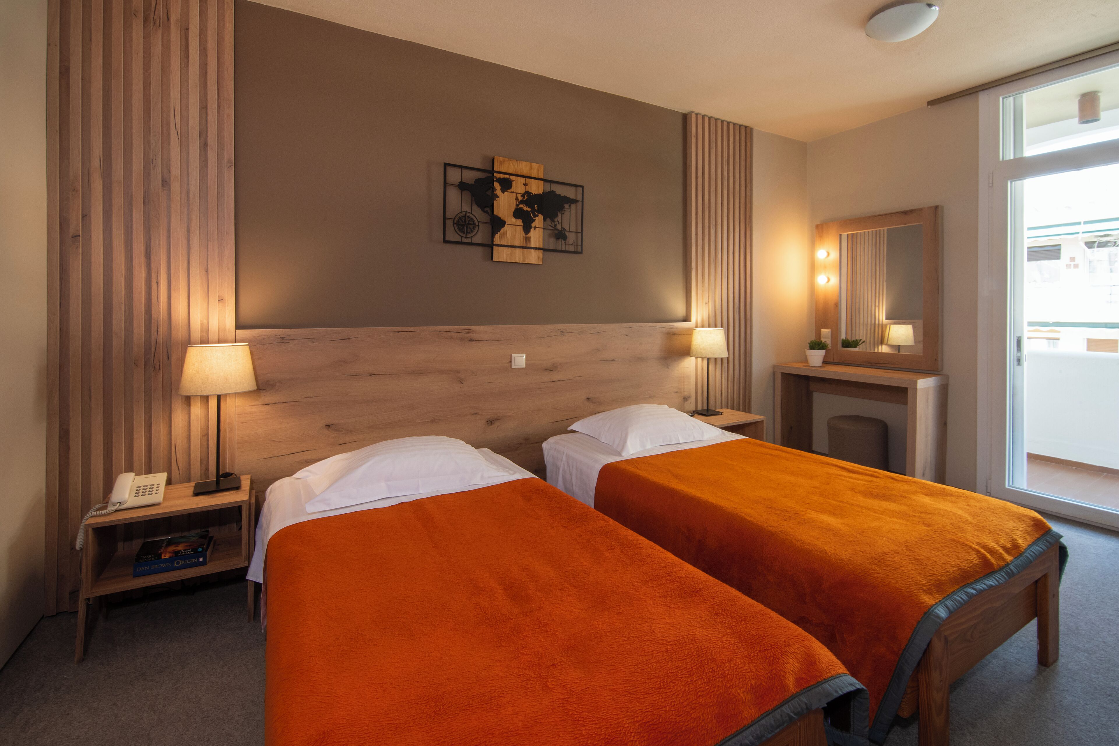 Habitación de hotel con dos camas, cabecero de madera, un cuadro con un mapa mundial, un espejo y una mesita de noche con lámpara y teléfono.
