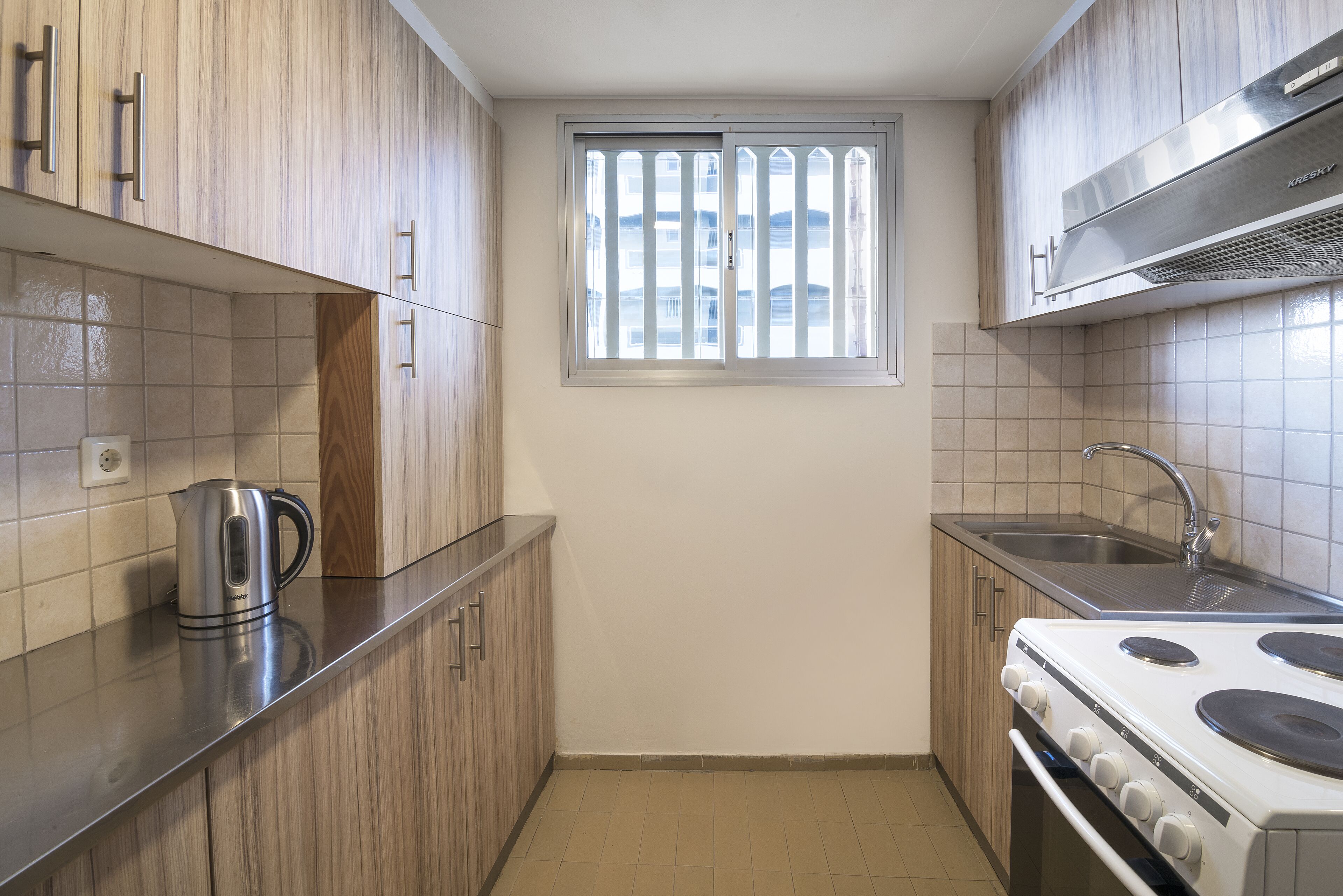 A compact kitchen with wooden cabinets, a stainless steel kettle, a white stove, and a window with bars.