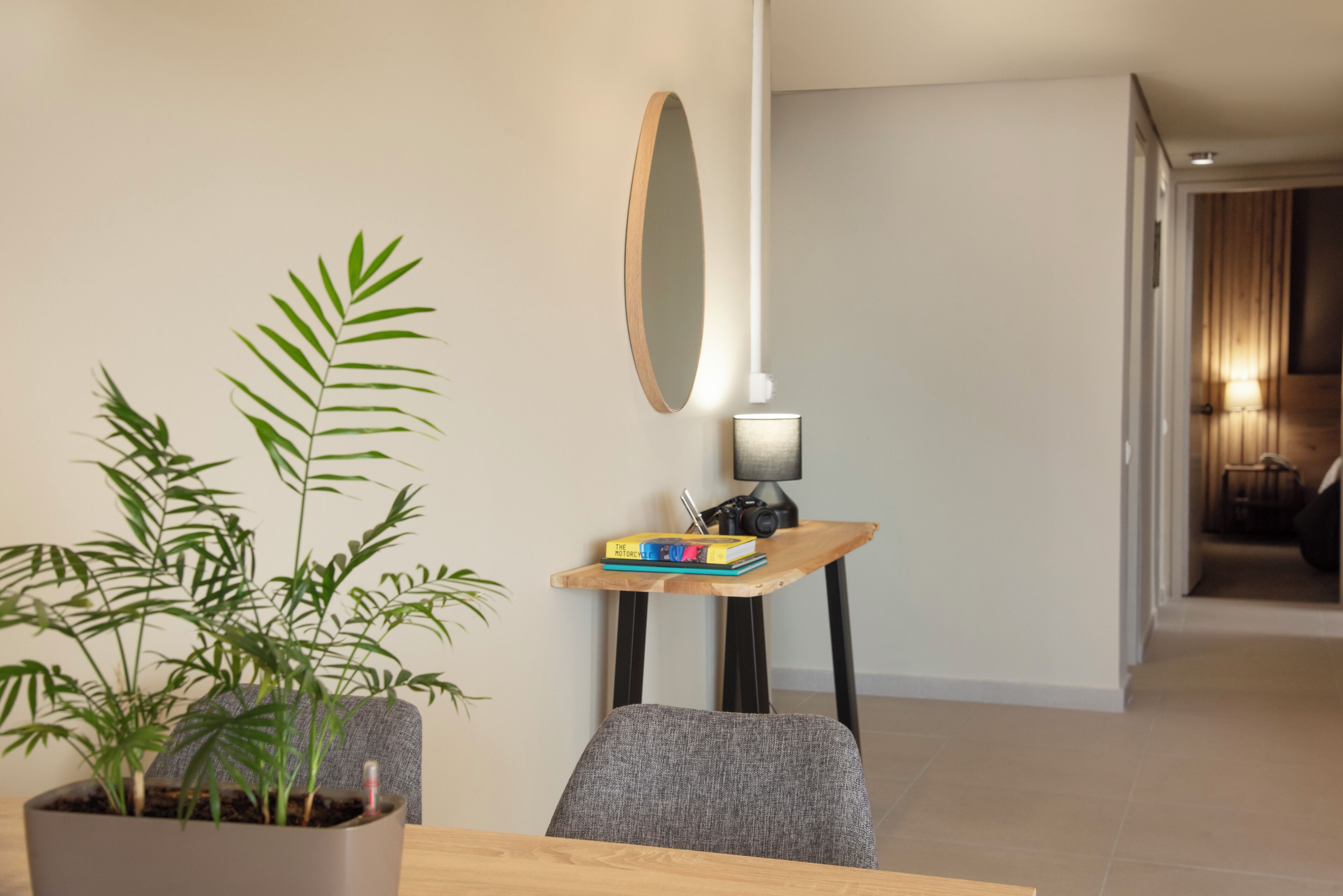 Un comedor moderno con una mesa de madera, una planta en maceta, una pila de libros, una lámpara pequeña y un espejo montado en la pared.