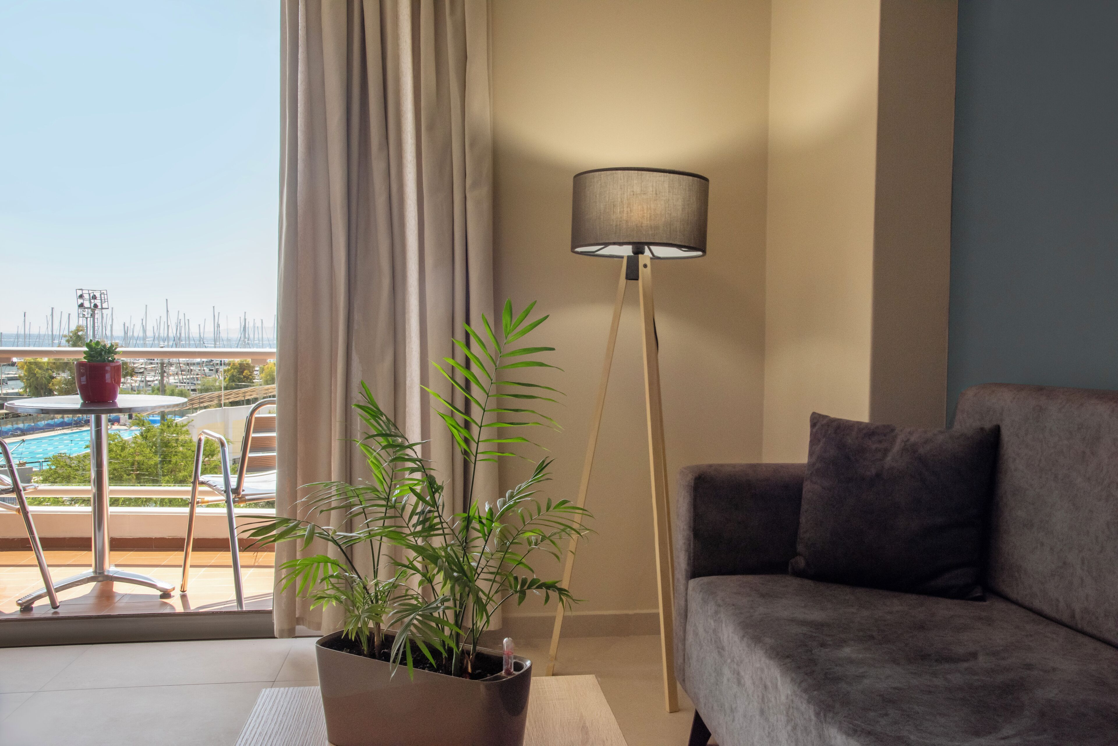 Un soggiorno moderno con un divano grigio, una lampada da terra e una pianta in vaso. Si intravede un balcone con piscina e mobili da esterno attraverso la finestra.