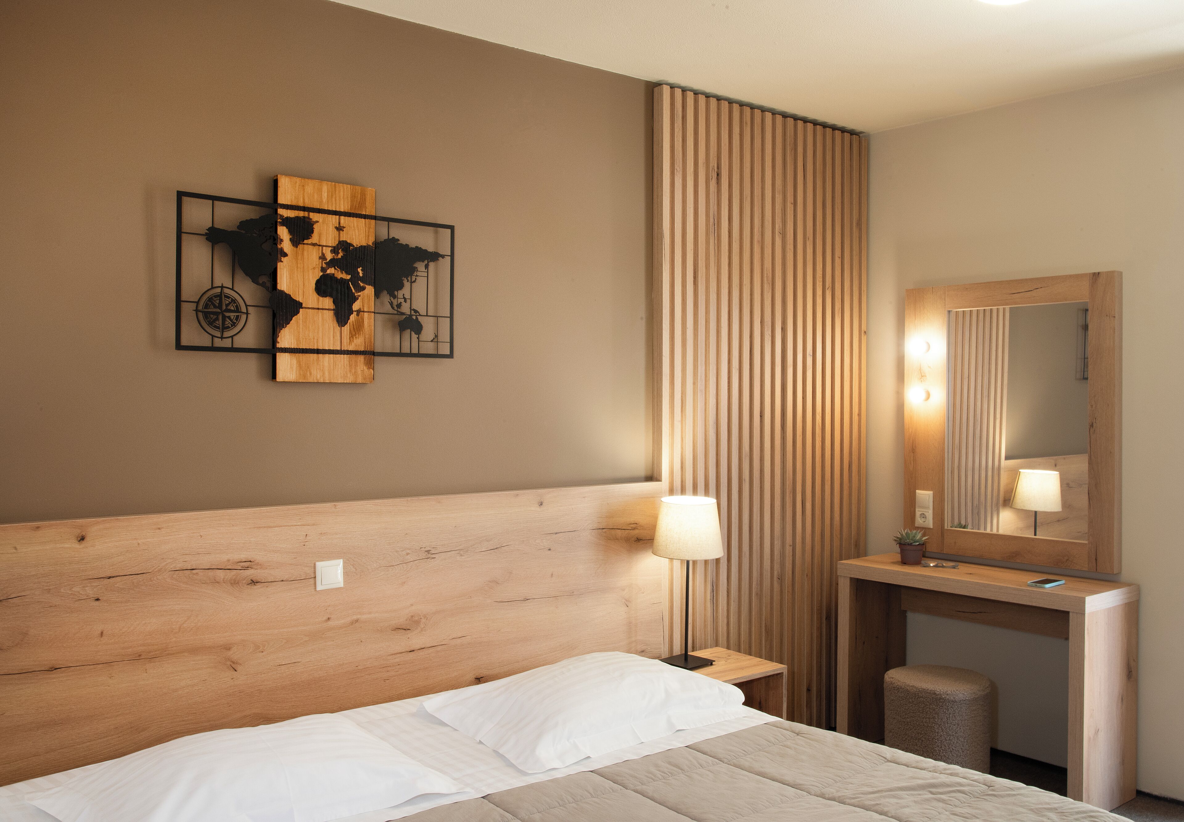 Une chambre d’hôtel avec une tête de lit en bois, un lit avec du linge de maison blanc, une table de chevet avec une lampe, un miroir et une œuvre d’art représentant une carte du monde accrochée au mur.