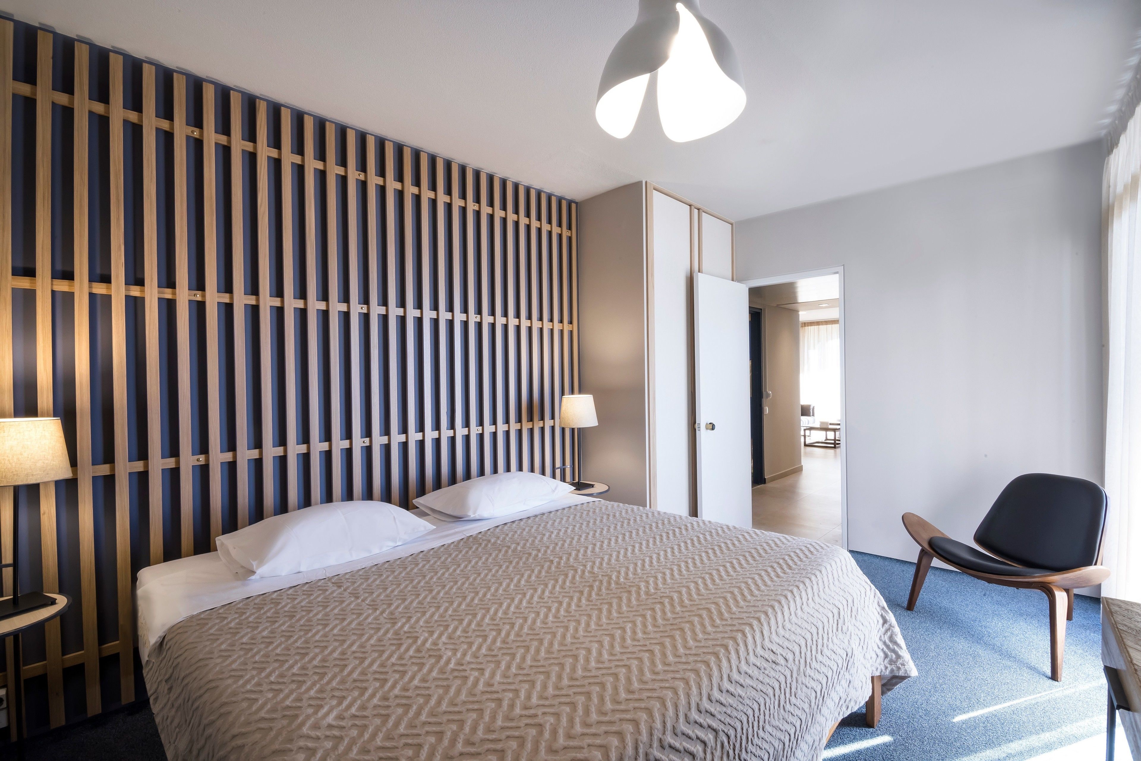 A modern hotel room with a large bed, a wooden slat headboard, a side table with a lamp, a black chair, and a view of the hallway.