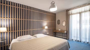 A hotel room with a large bed, a bedside table with a lamp, a desk with a chair, a wall with vertical wooden slats, and a window with sheer curtains.
