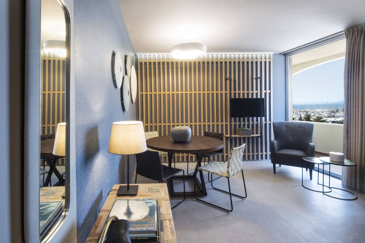 Moderni hotellihuone, jossa on ruokailutila, sohva, televisio ja näkymä kaupunkiin.