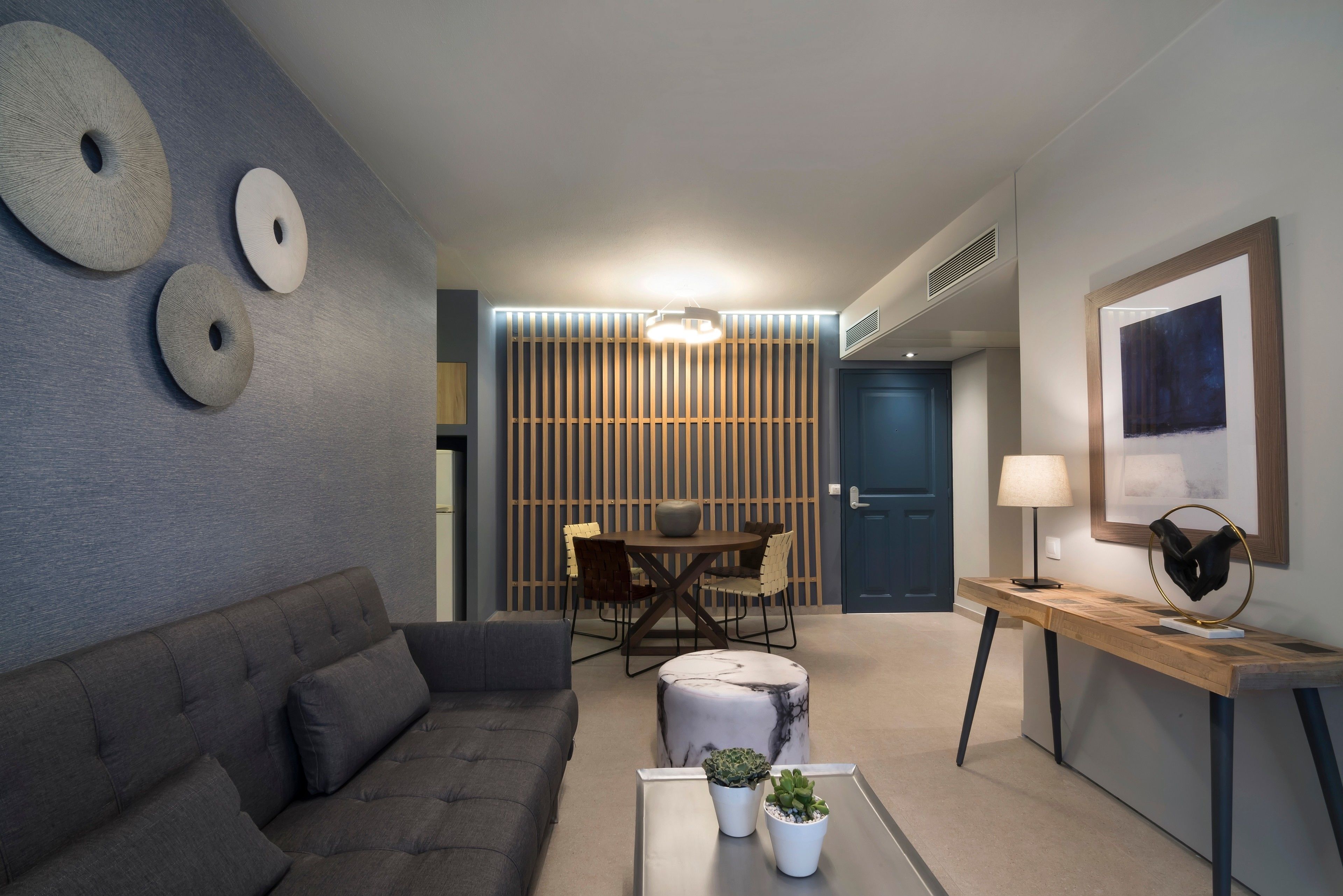 A modern living room with a grey sofa, a round coffee table with plants, a wooden dining table with chairs, and a blue door in the background.