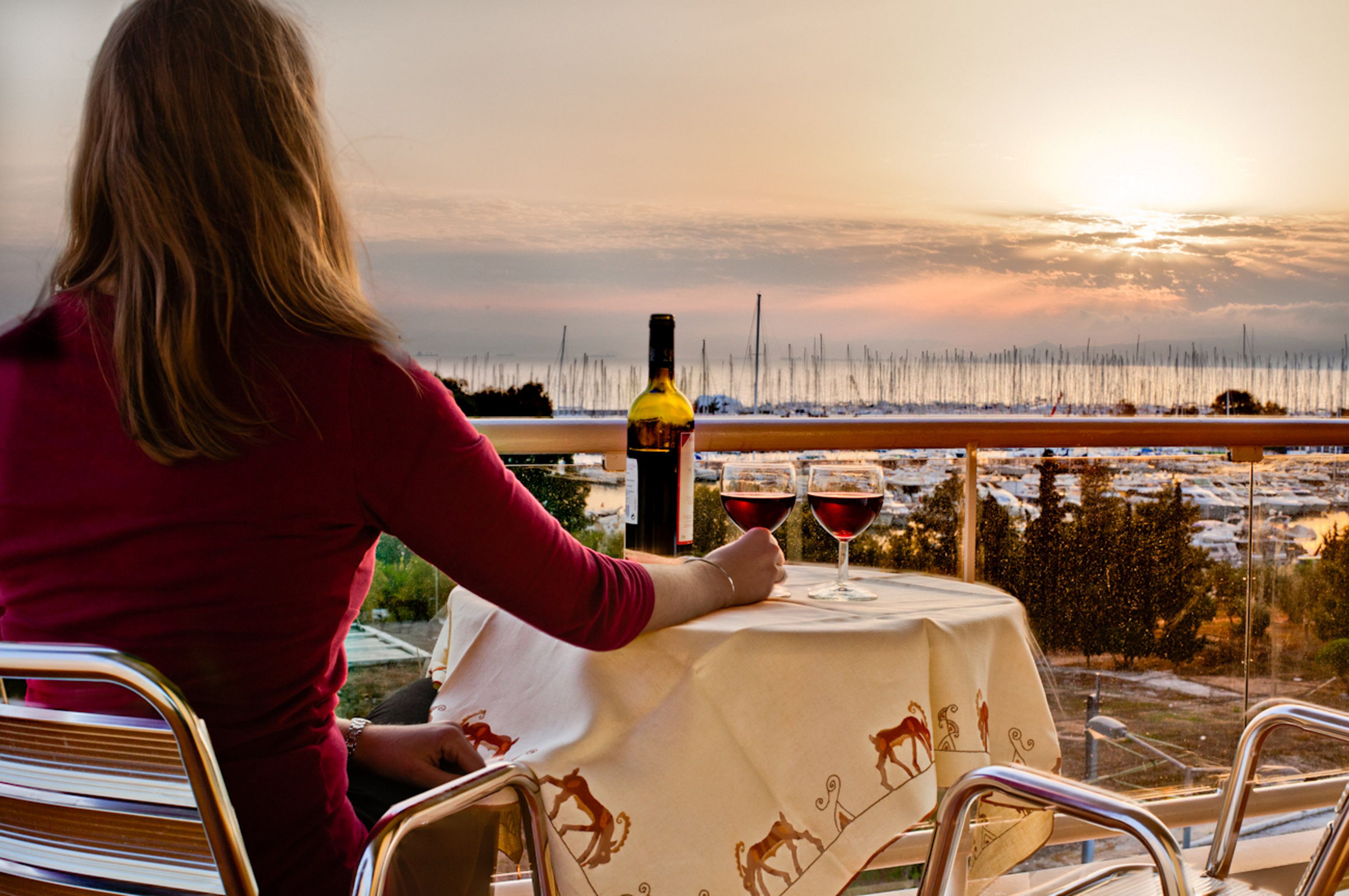 Una persona sentada en una mesa al aire libre con una botella de vino y dos copas, con vistas a un paisaje nevado al atardecer.