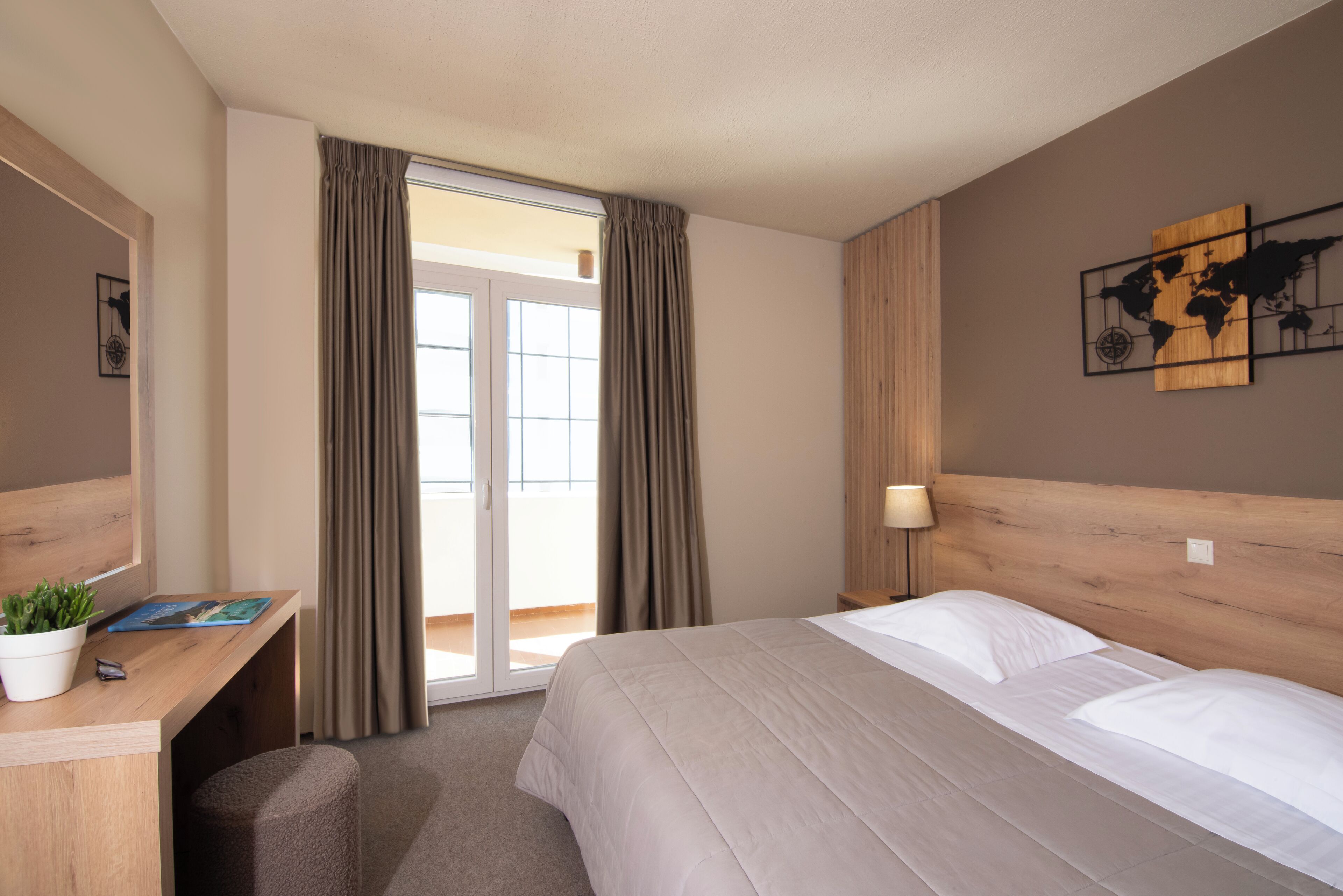 Une chambre d’hôtel avec un lit, un bureau agrémenté d’une plante, une œuvre d’art représentant une carte du monde et une vue sur l’extérieur, visible à travers une porte vitrée.