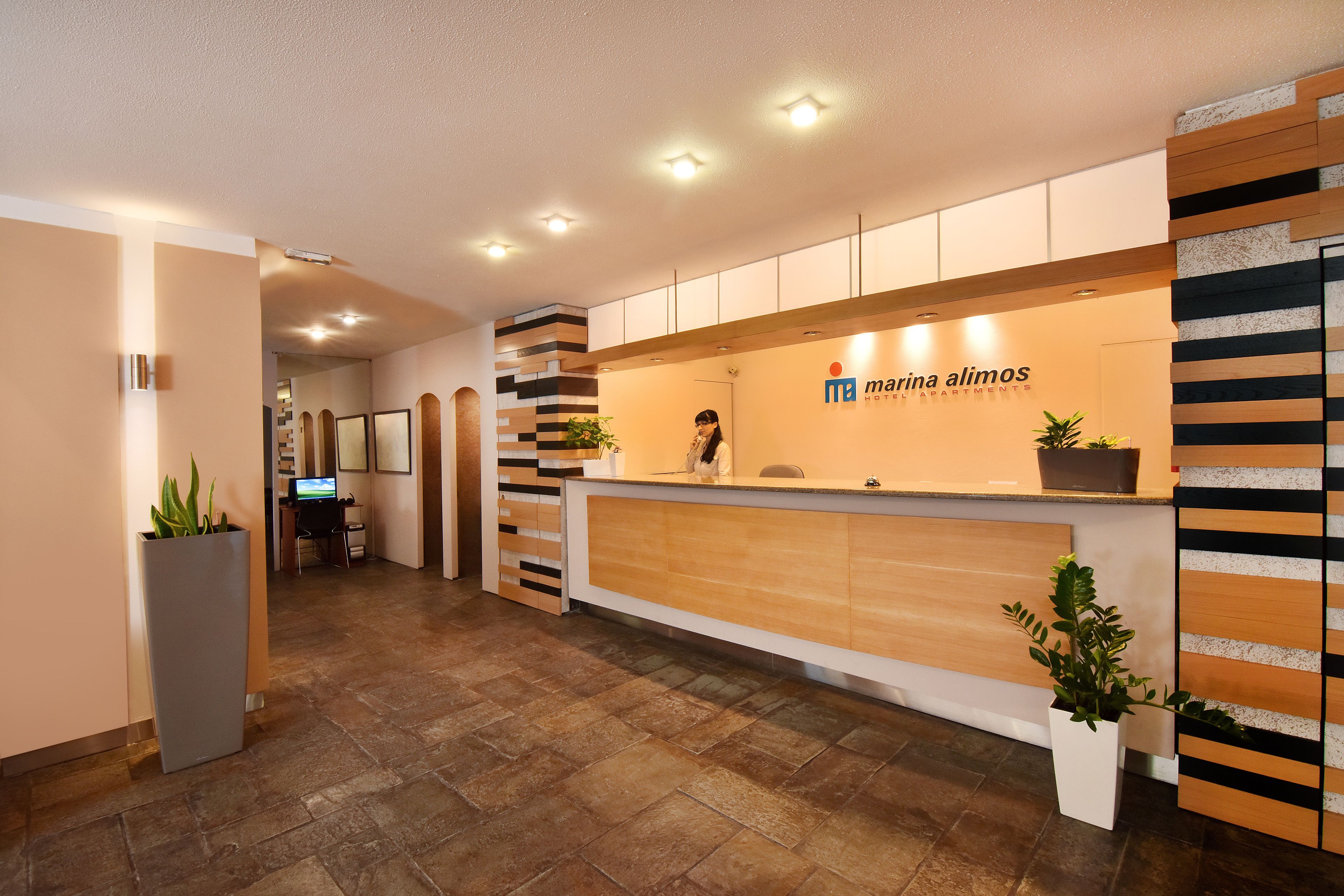 A reception area with a reception desk, potted plants, and a sign for Marina Alimos Hotel Apartments.