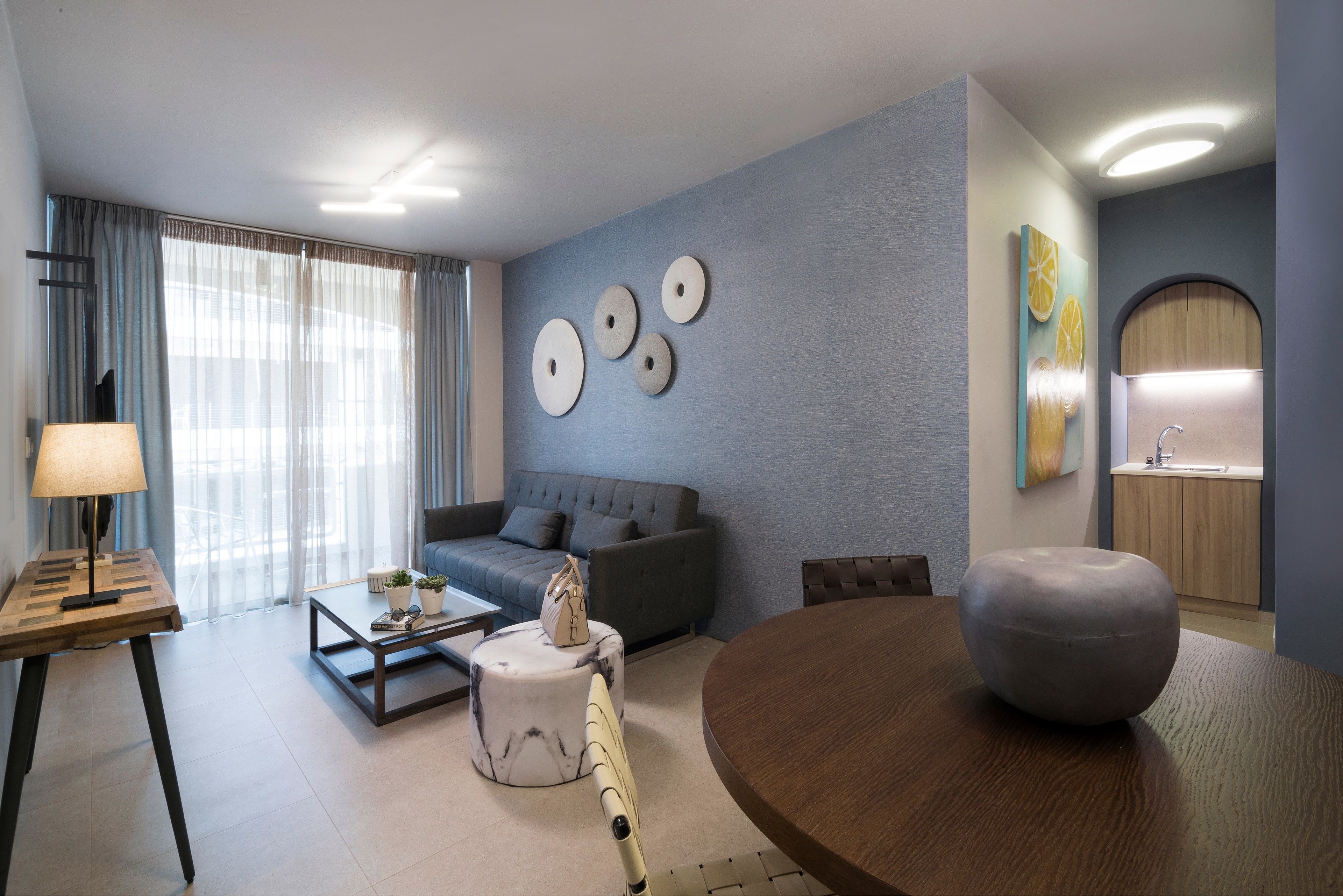 A modern living room with a grey sofa, a round wooden table, a coffee table, and a lamp. There are decorative plates on the wall and a kitchen area in the background.
