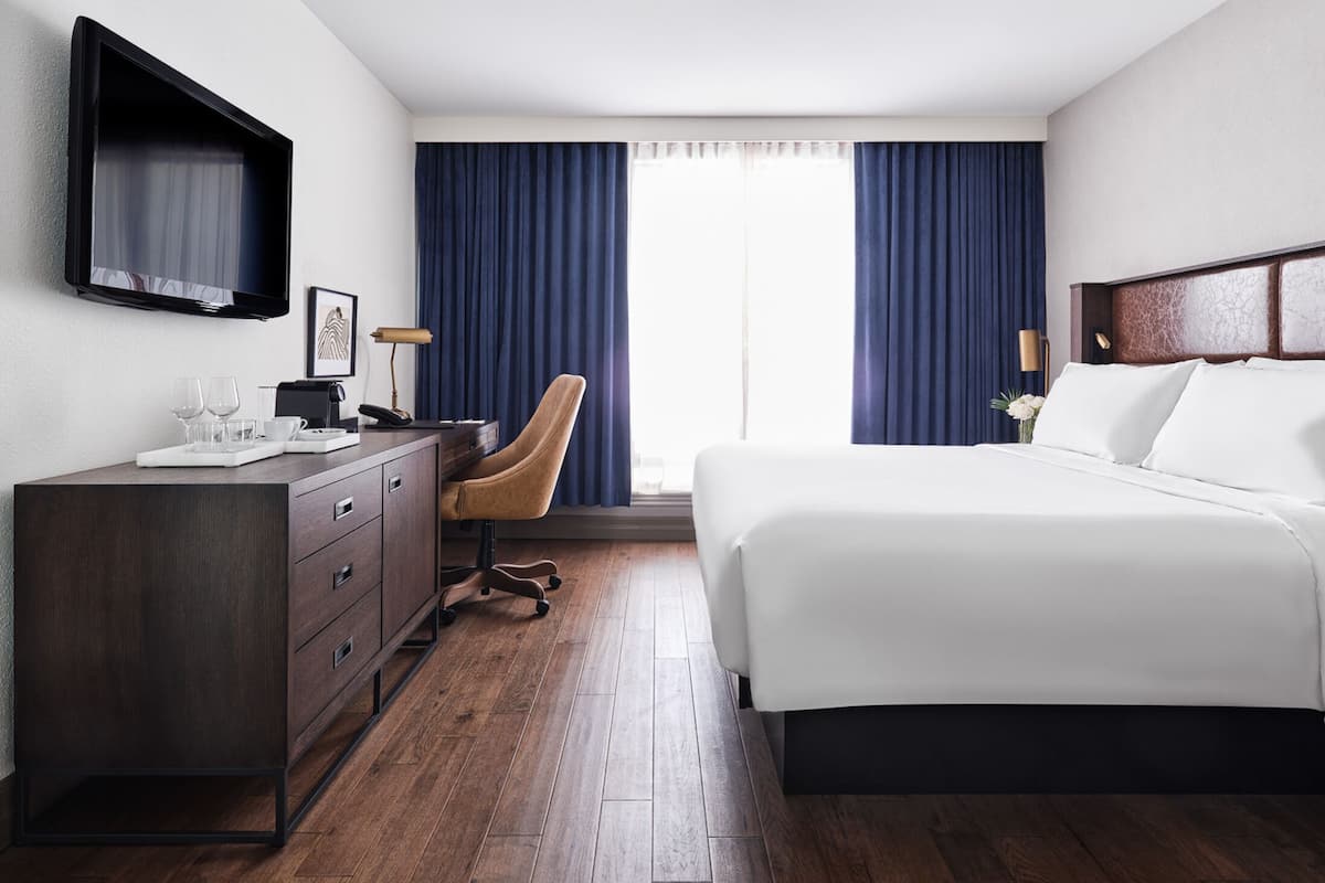 A hotel room with a bed, a desk with a chair, a TV, and a window with curtains.