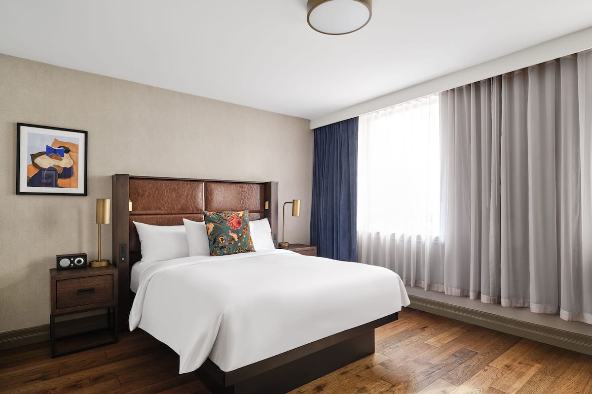 A hotel room with a large bed, a nightstand with a radio, a framed picture on the wall, and a window with curtains.