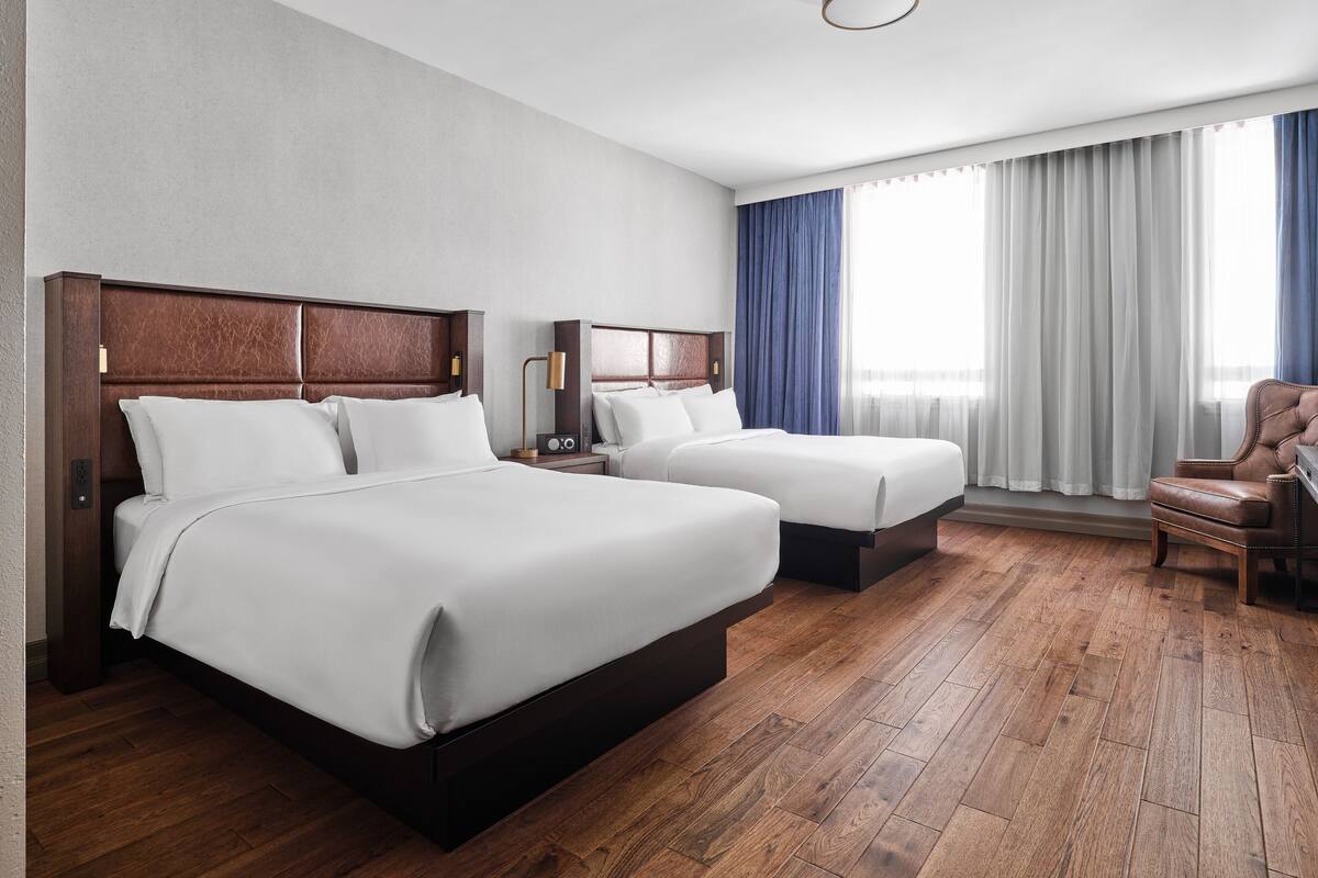 A hotel room with two beds, a wooden floor, a ceiling light, and a window with curtains.