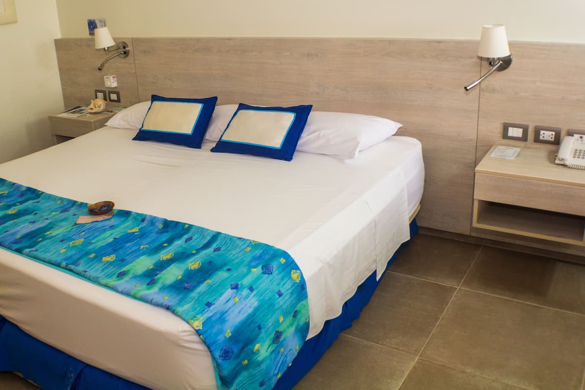 Quarto de hotel com cama grande, duas almofadas, mesa de cabeceira com abajur e telefone.