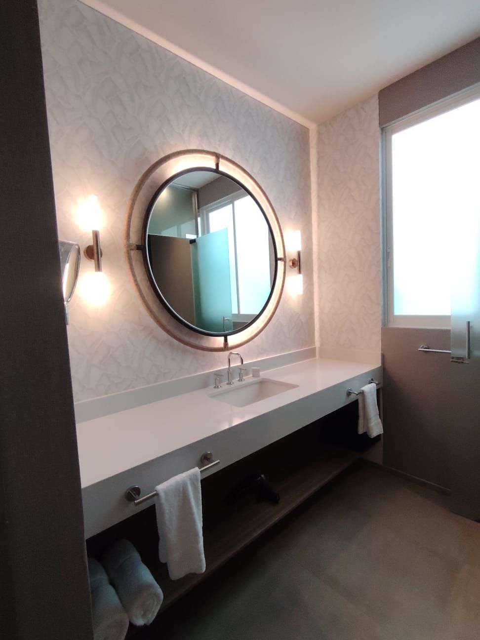 Un baño con un espejo redondo, dos lavamanos y un perchero para toallas.