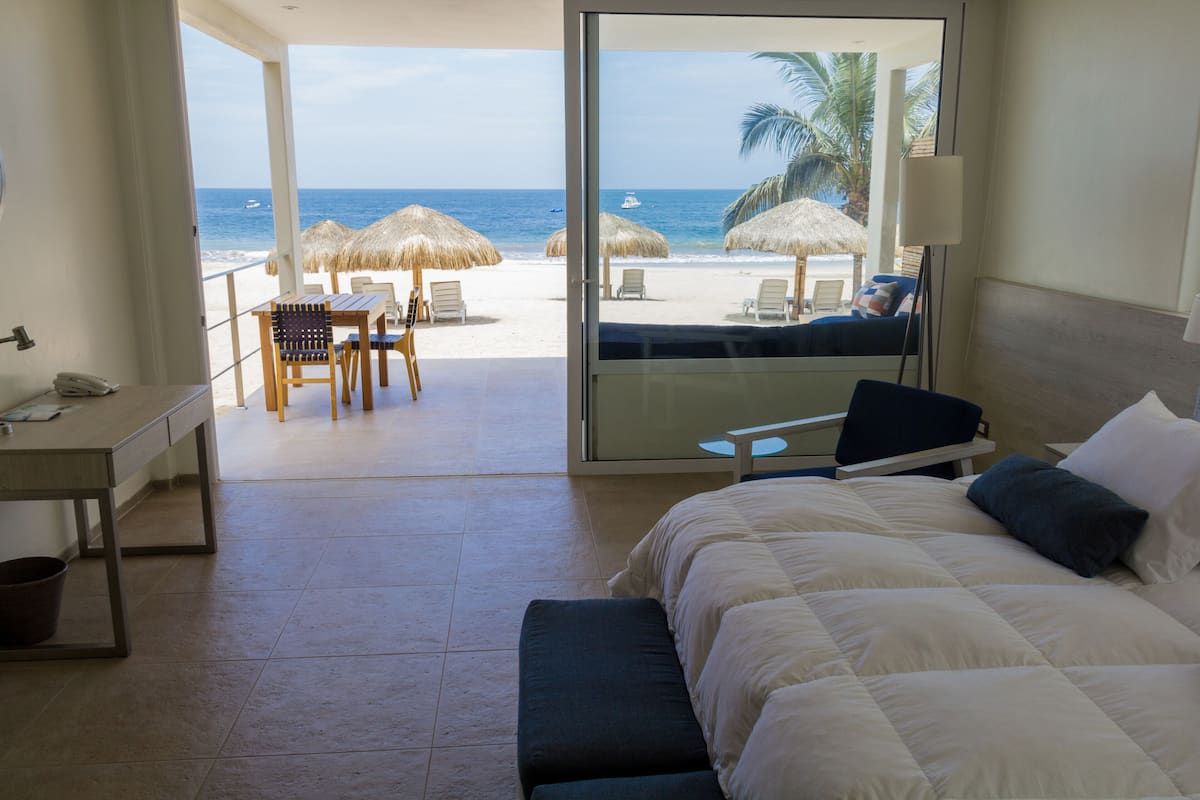 Quarto de hotel com cama, escrivaninha, cadeira e vista para a praia.