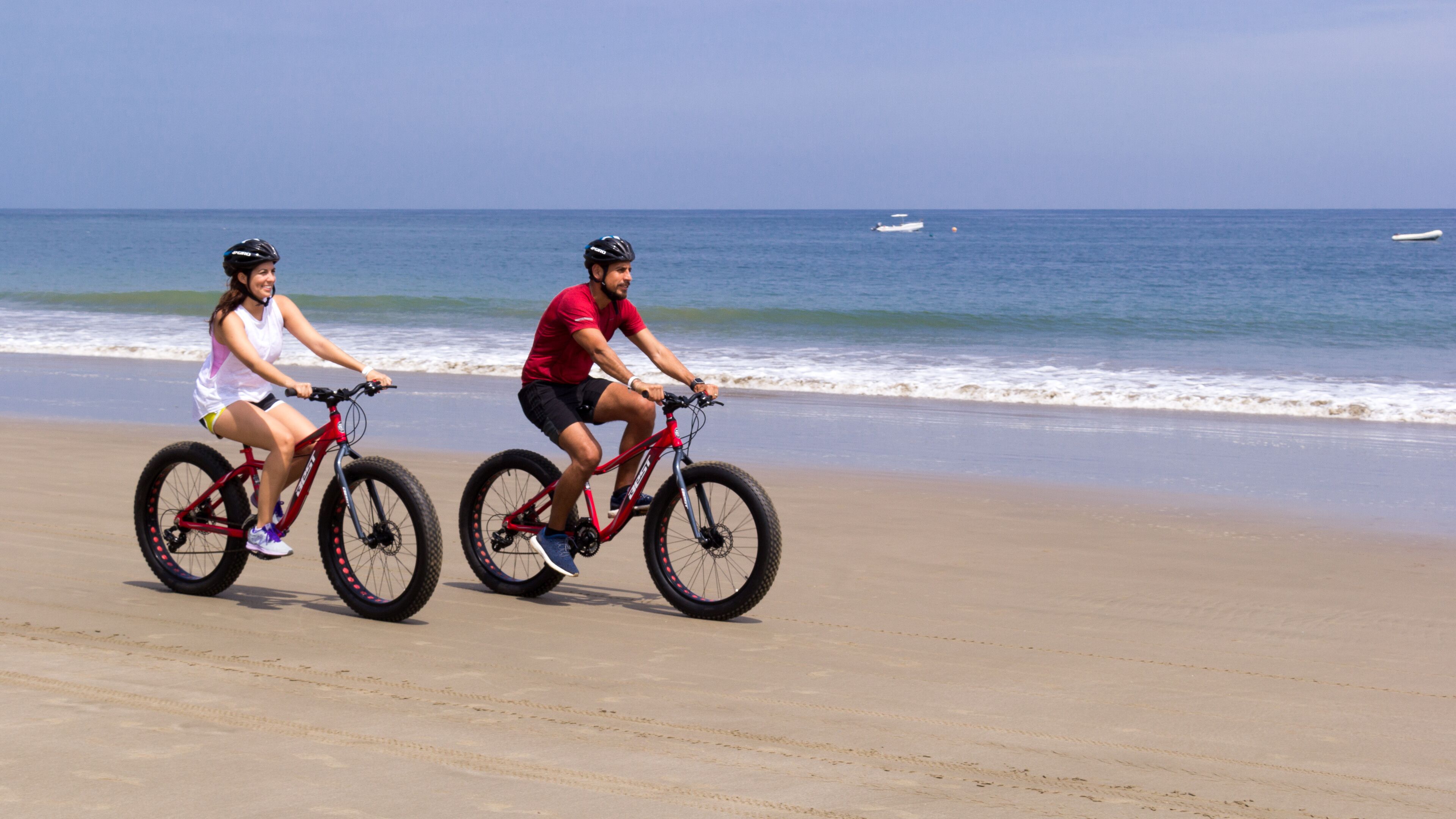 Dos personas paseando en bicicleta sobre una playa arenosa, con el océano al fondo.