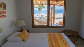 A bedroom with a bed, a nightstand, a lamp, a window with a view of the beach, and a wooden bench.