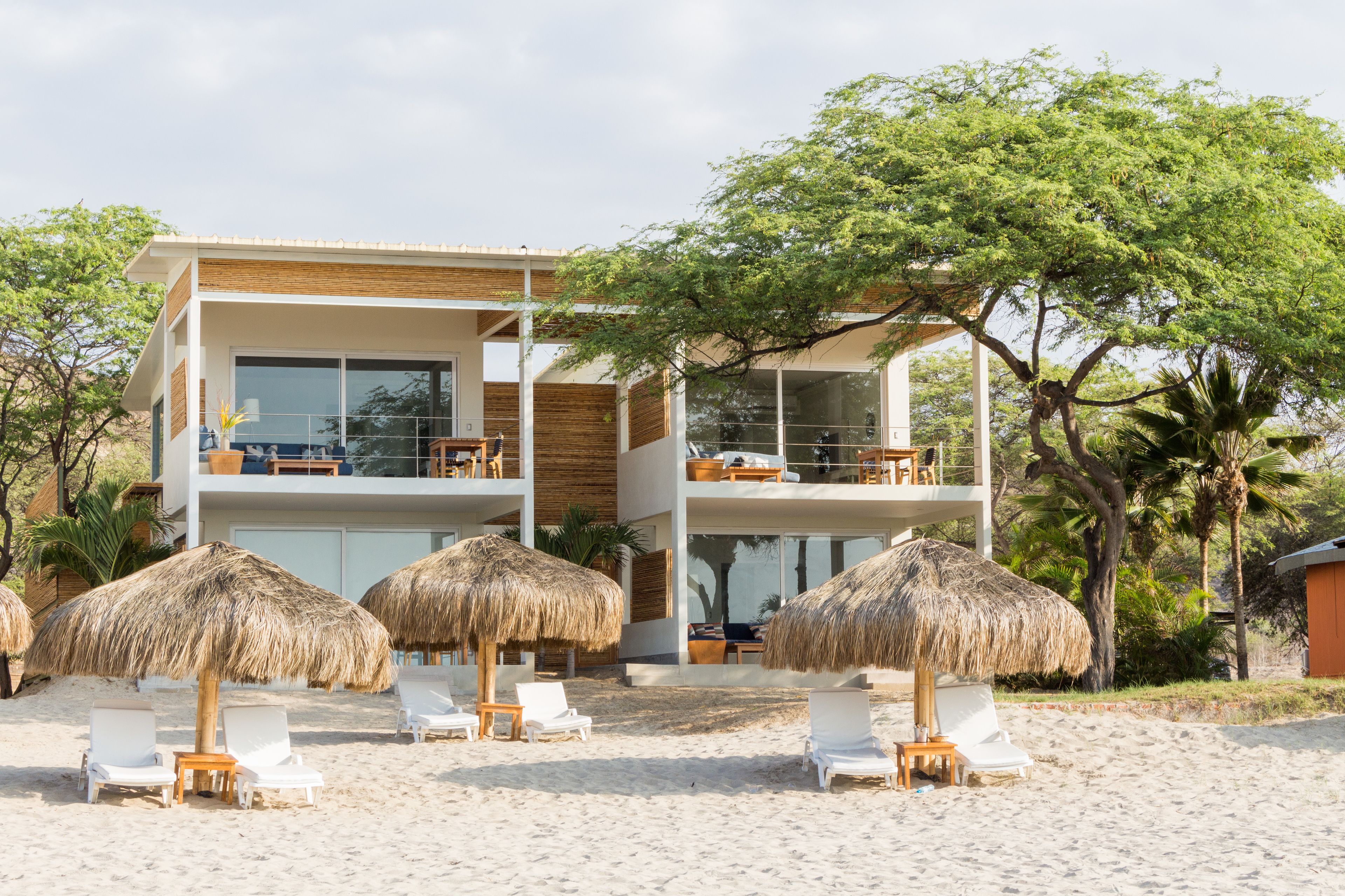 Una casa moderna frente a la playa con dos balcones, sillones blancos y sombrillas de paja.