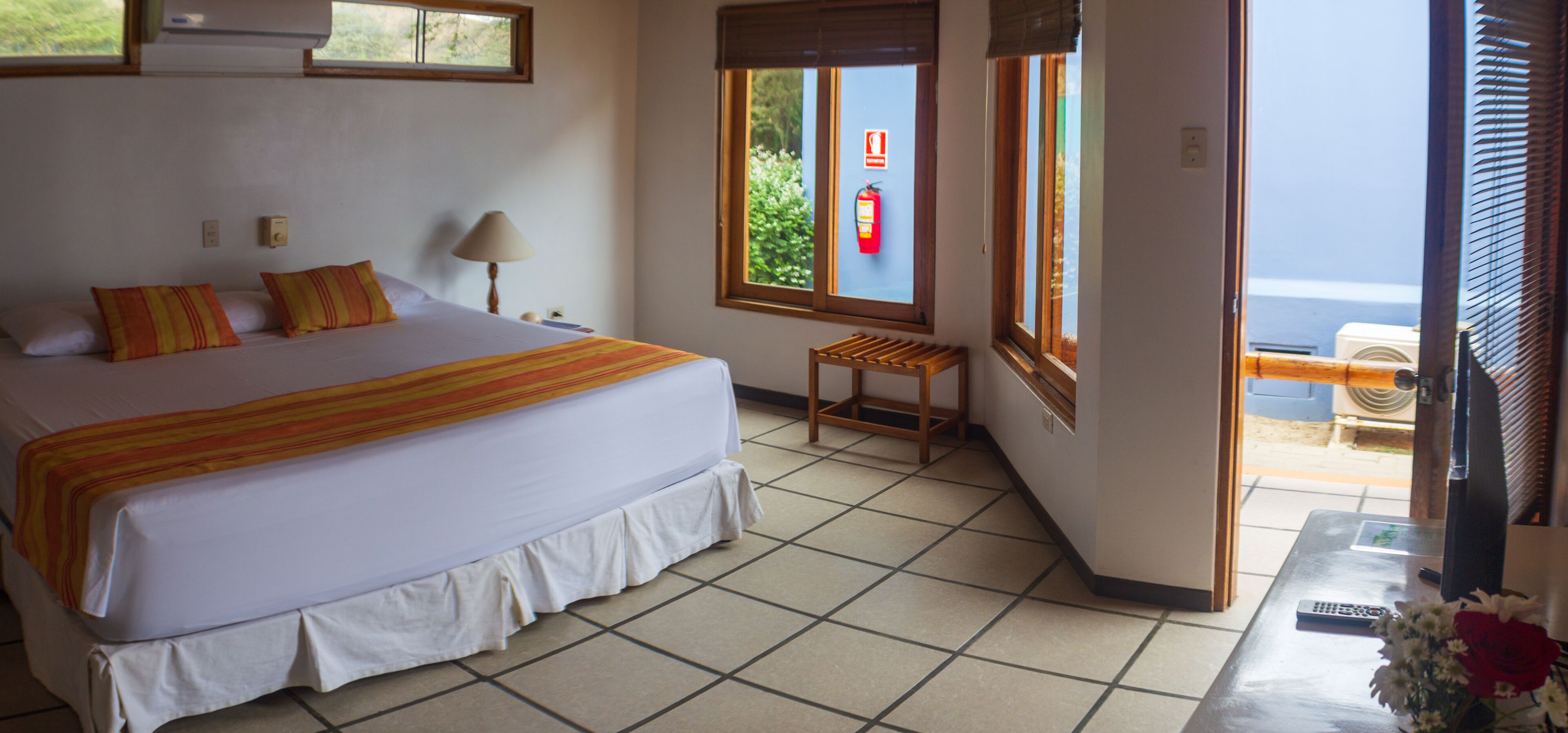 Habitación de hotel con cama, mesita de noche, lámpara, ventana con vista y una pequeña mesa con un jarrón de flores.