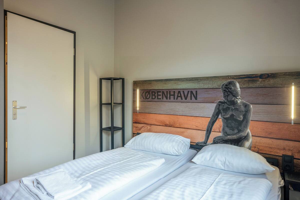 A bedroom with a bed, a wooden headboard with the text "KÖBENHAVN" and a statue of a seated figure.