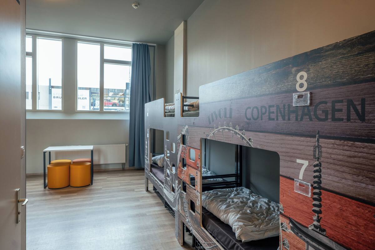 A room with bunk beds, a table with two stools, and a window with a view of buildings.
