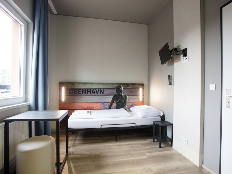 Quarto com cama, mesa, cadeira, televisão fixada na parede e janela com cortinas.