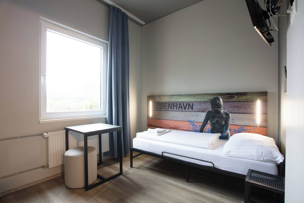 A modern hotel room with a bed, a small table, a radiator, and a window with curtains.
